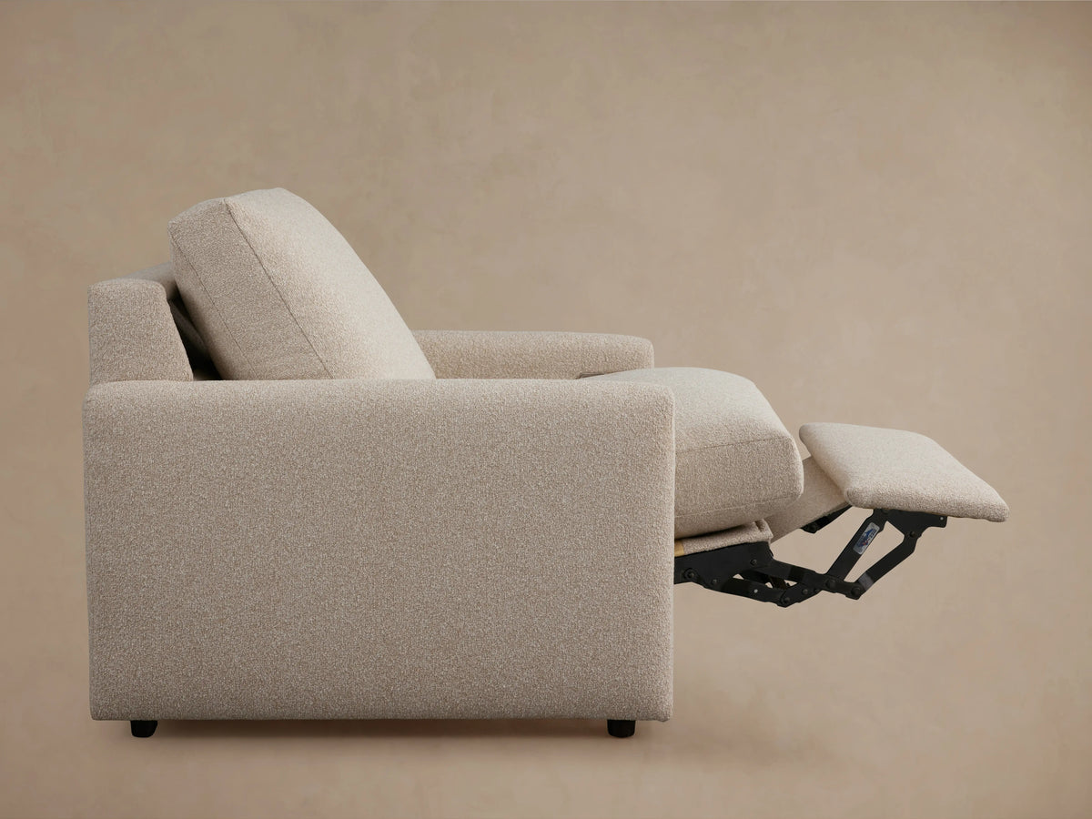 Kipton Motion Recliner close-up shows beige fabric texture, slim armrest with metal button for reclining, and cushion detail.