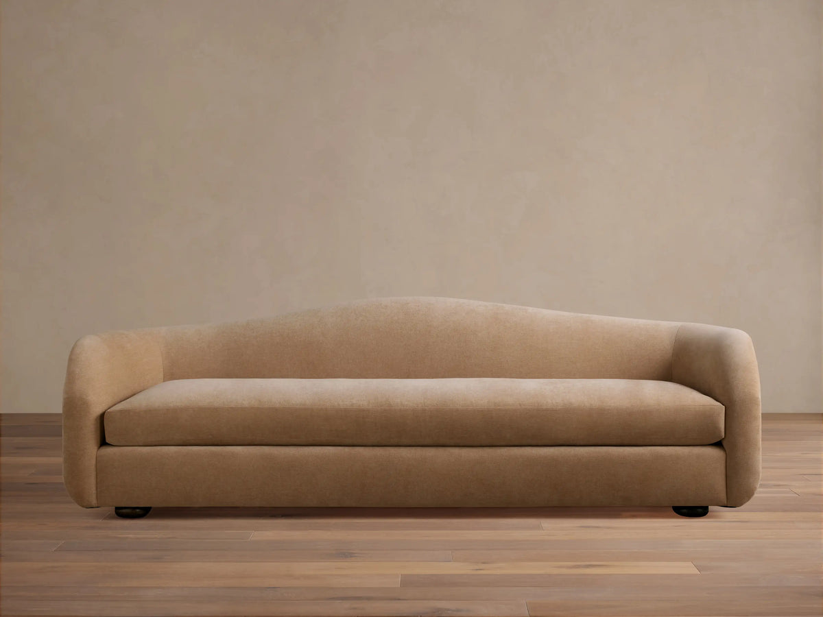 Ashlyn Sofa in warm tan, front view. Modern, minimalist design with a gently curved backrest and integrated arms on a wooden floor.