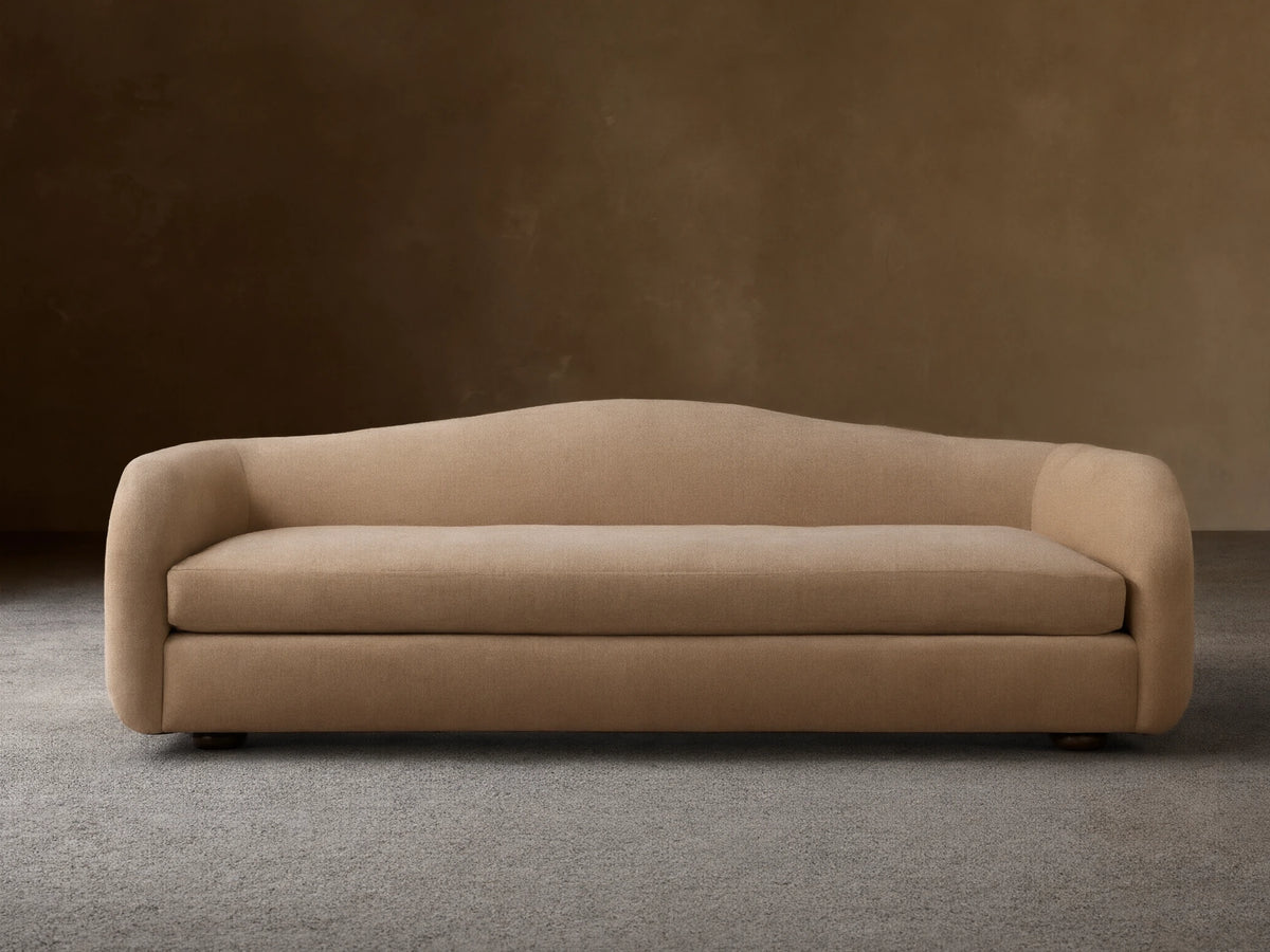 Ashlyn Sofa in warm tan, front view. Modern, minimalist design with a gently curved backrest and integrated arms on a wooden floor.
