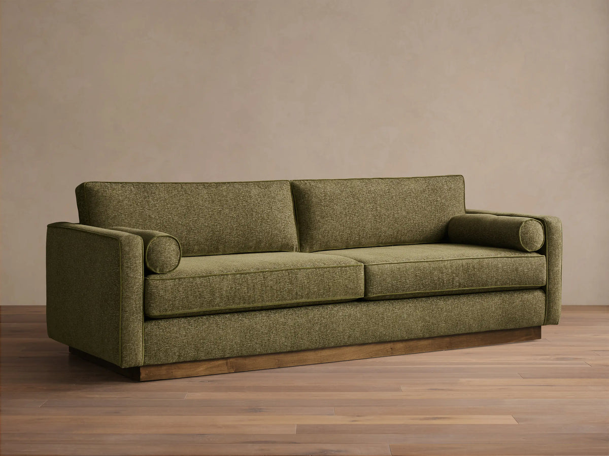 Front view of the Plush Olive Munroe Sofa, featuring two cylindrical arm pillows and a contrasting wooden base.