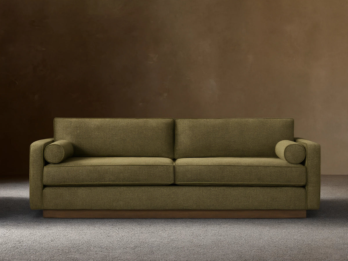 Front view of the Plush Olive Munroe Sofa, featuring two cylindrical arm pillows and a contrasting wooden base.