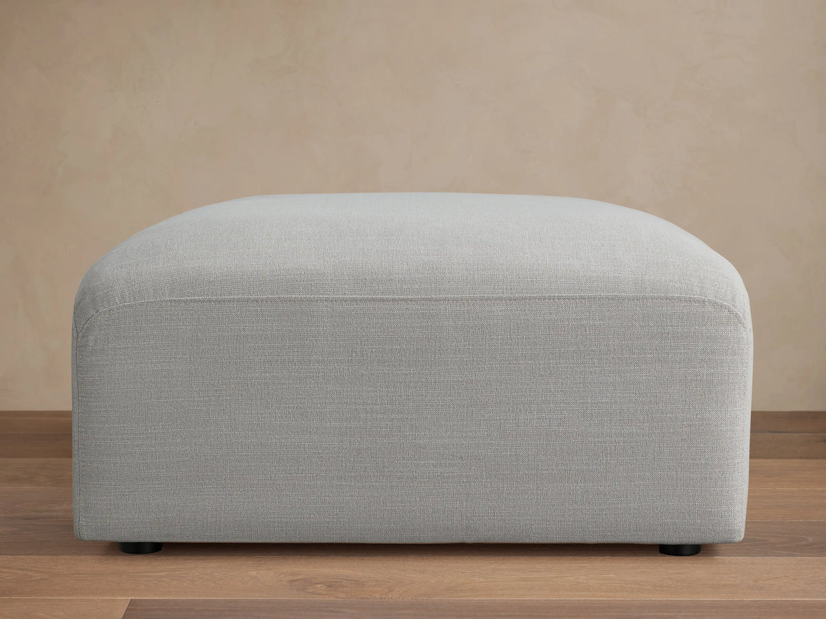Duncan Ottoman, light grey upholstered fabric with subtle texture and rounded edges, on a wooden floor with a plain beige wall.