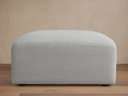 Duncan Ottoman, light grey upholstered fabric with subtle texture and rounded edges, on a wooden floor with a plain beige wall.