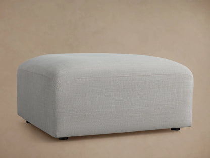 Duncan Ottoman, light grey upholstered fabric with subtle texture and rounded edges, on a wooden floor with a plain beige wall.