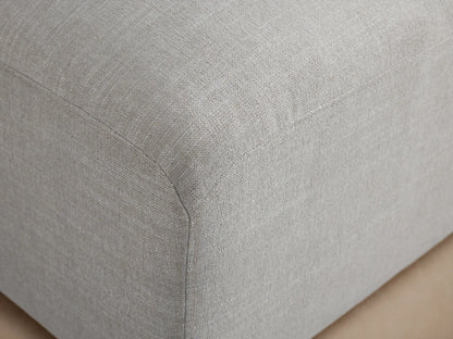 Duncan Ottoman, rectangular with rounded top, upholstered in light gray woven fabric, shown at an angle on small black feet.