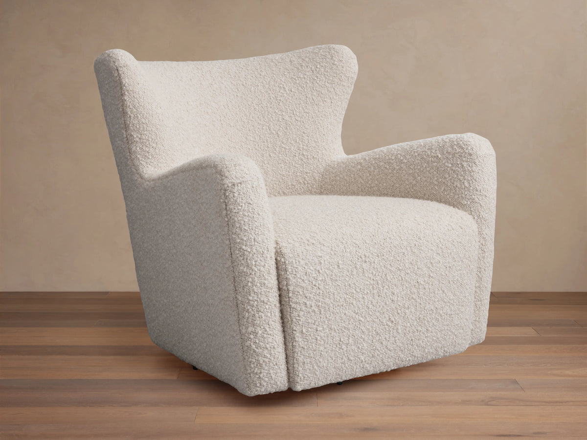 A cream-colored Sasha Swivel Chair with a textured fabric. It features a rounded backrest and armrests, set on a wooden floor.