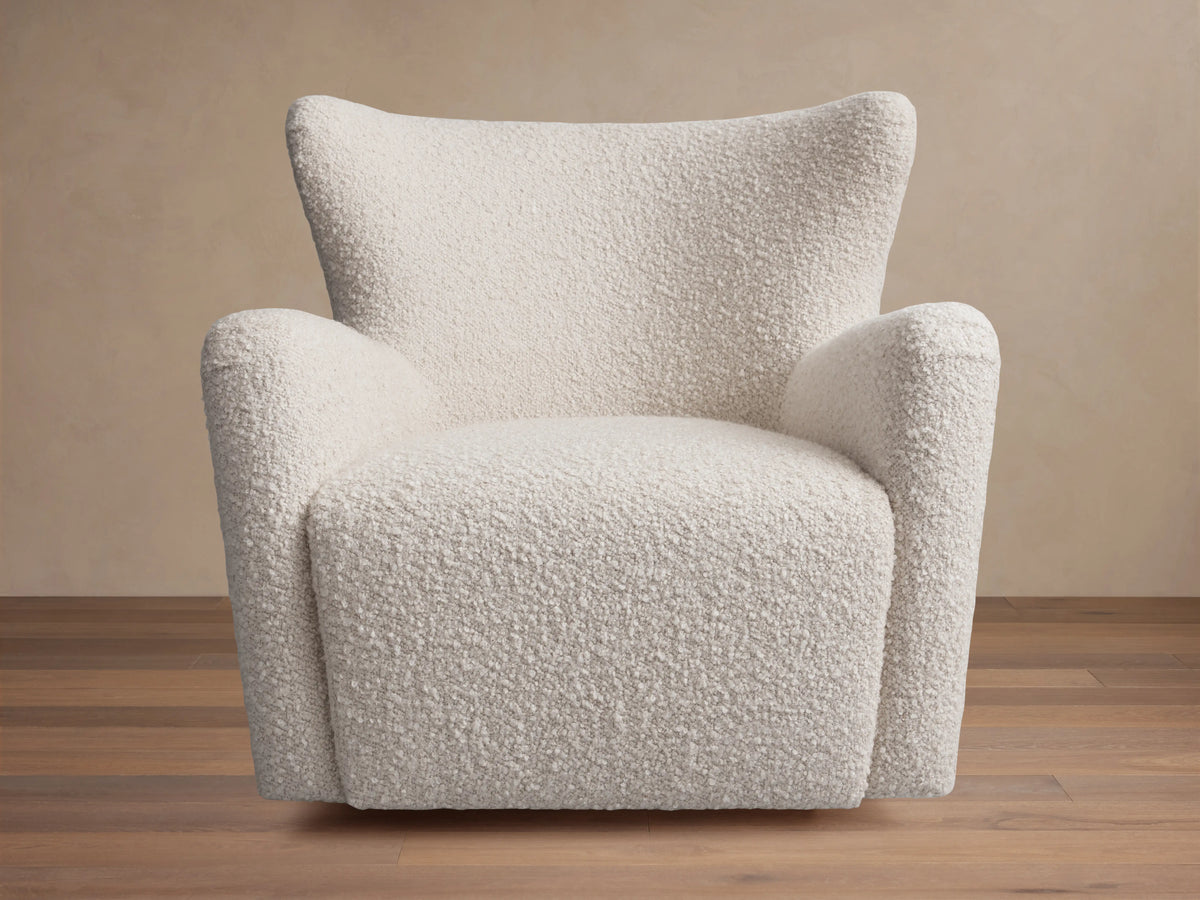 A cream-colored Sasha Swivel Chair with a textured fabric. It features a rounded backrest and armrests, set on a wooden floor.