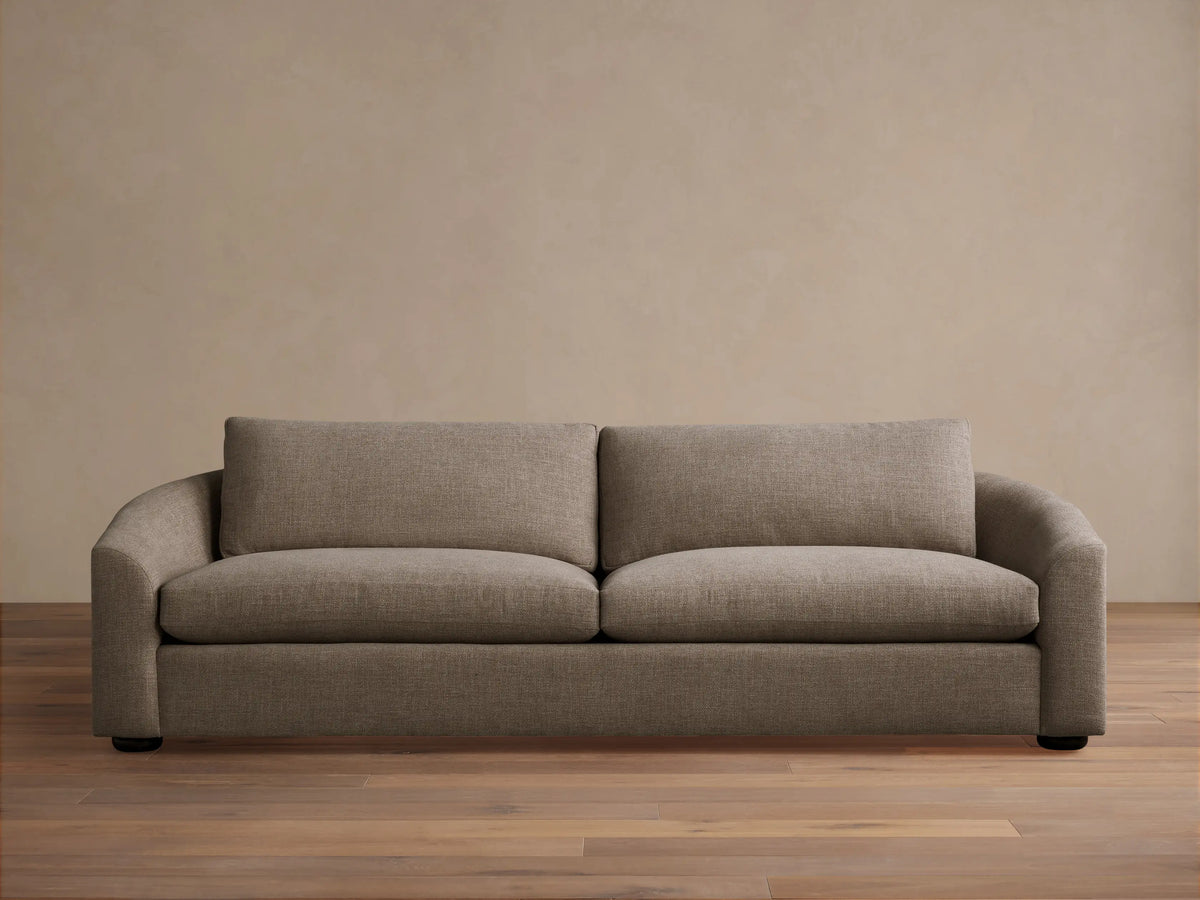 Avon Sofa in light brown textured fabric, front view, with two seat & back cushions, curved arms, on wood floor against beige wall.