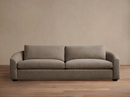 Avon Sofa in light brown textured fabric, front view, with two seat & back cushions, curved arms, on wood floor against beige wall.