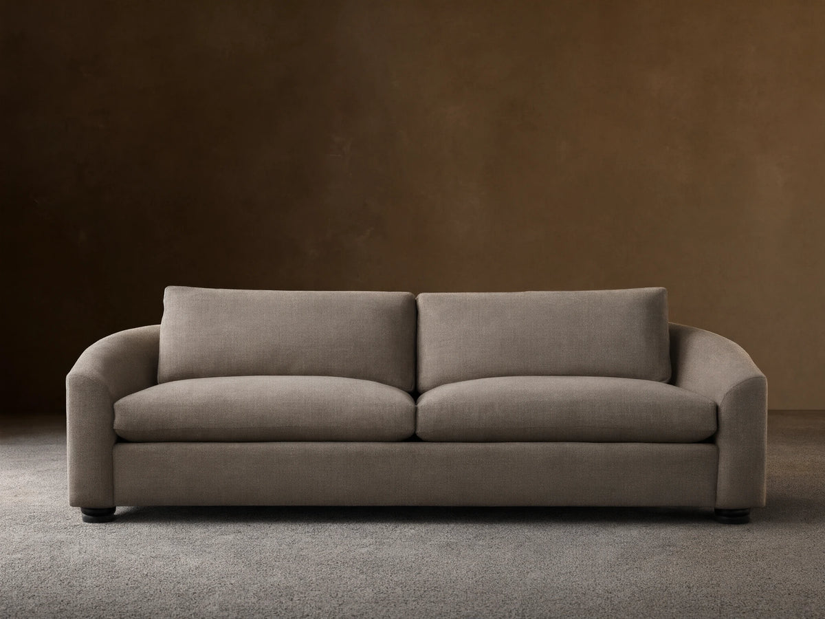 Avon Sofa in light brown textured fabric, front view, with two seat & back cushions, curved arms, on wood floor against beige wall.
