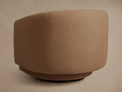 Round, light brown ottoman, upholstered in fine textured fabric, seen from the back. It sits on a dark, subtle circular base.