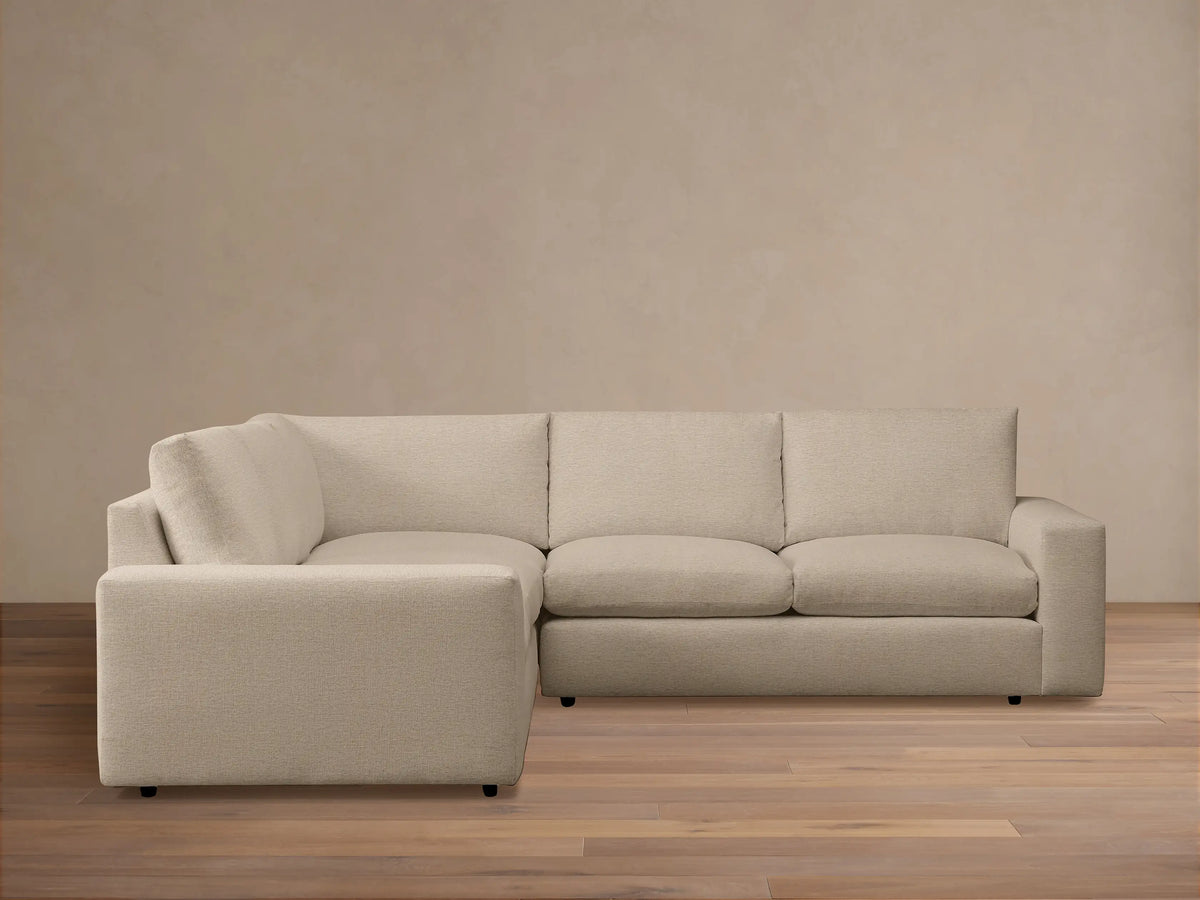 Kipton Wide Arm Three-Piece Corner Sectional in beige fabric with wide armrests, positioned in a minimalist room on a wooden floor.