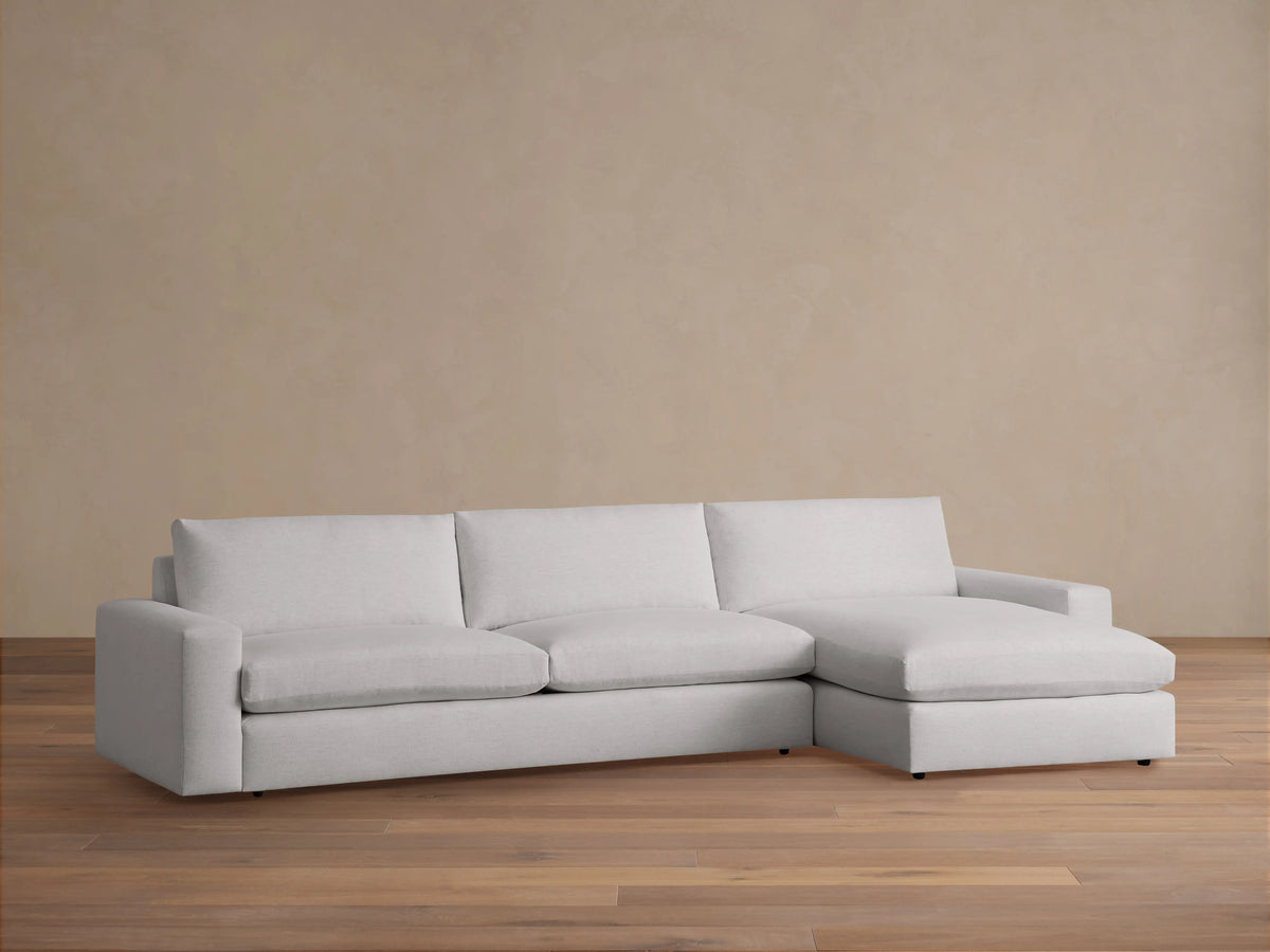 Wide arm two-piece chaise sectional in light gray fabric positioned against a beige wall on wooden flooring. The chaise is on the right.