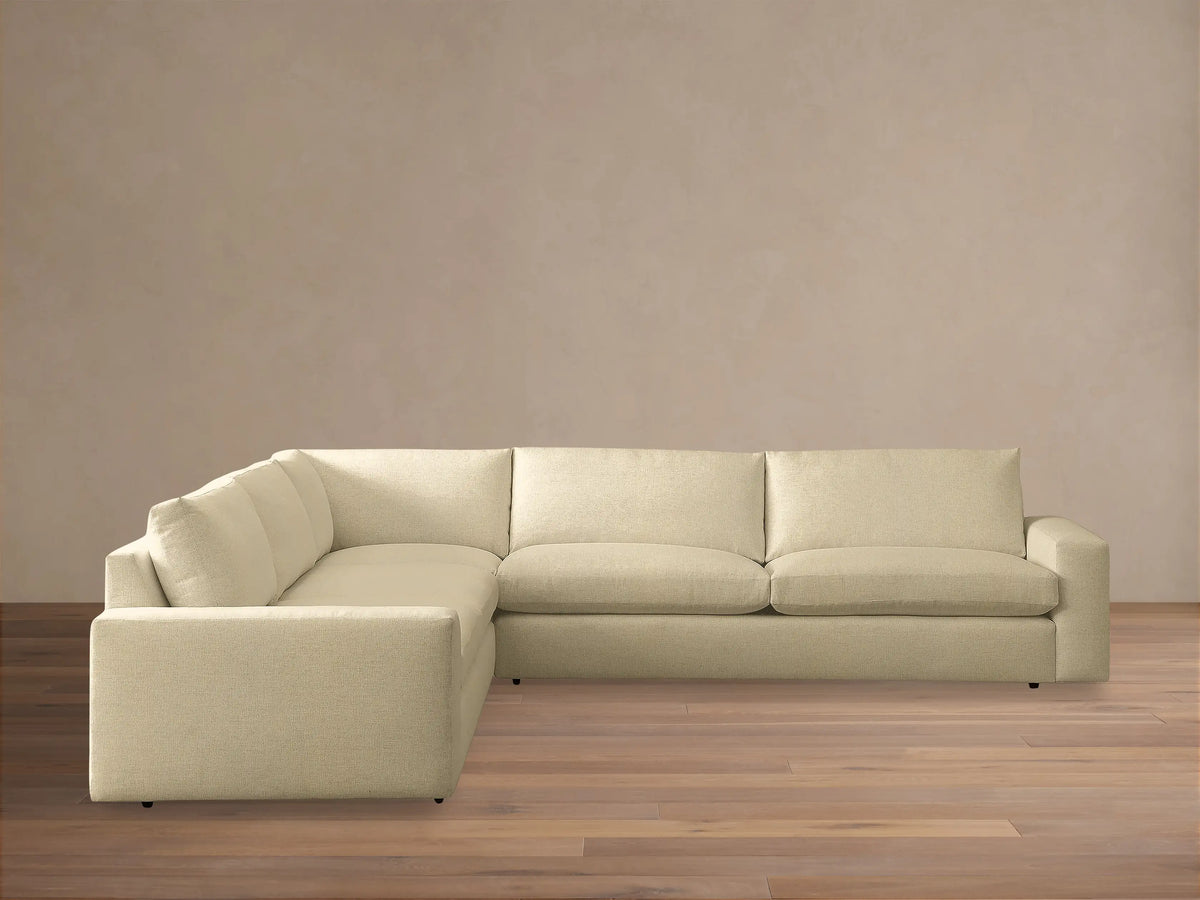 Kipton Wide Arm Three-Piece Corner Sectional, light beige L-shaped sofa on a wood floor, against a tan wall.