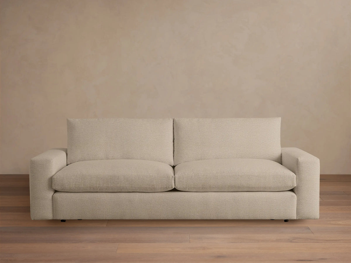 Kipton Wide Arm Sofa in beige fabric, featuring a minimalist design with wide armrests and cushioned backrests, set on a wooden floor.