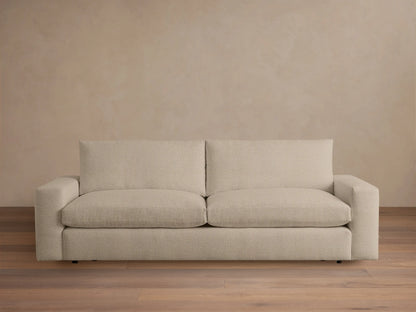 Kipton Wide Arm Sofa in beige fabric, featuring a minimalist design with wide armrests and cushioned backrests, set on a wooden floor.