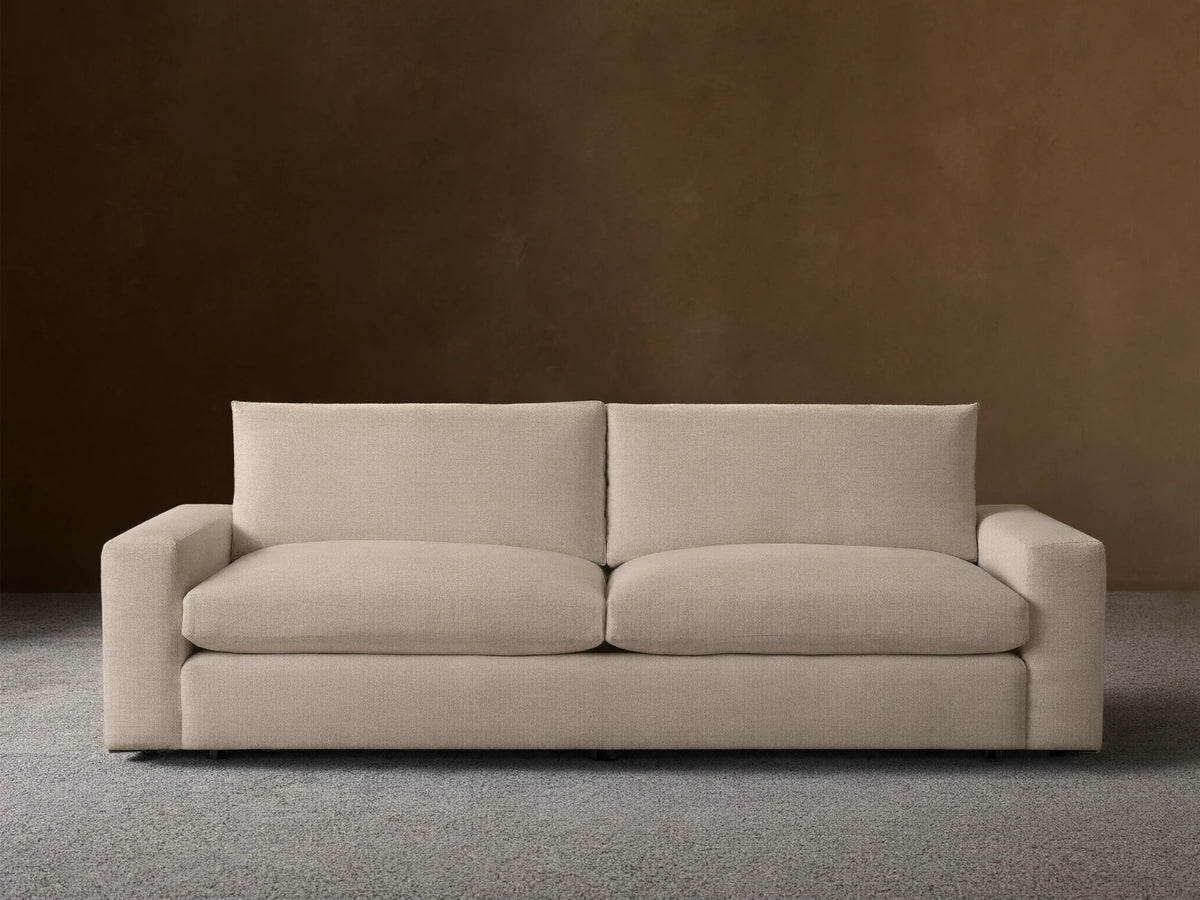 Kipton Wide Arm Sofa in beige fabric, featuring a minimalist design with wide armrests and cushioned backrests, set on a wooden floor.
