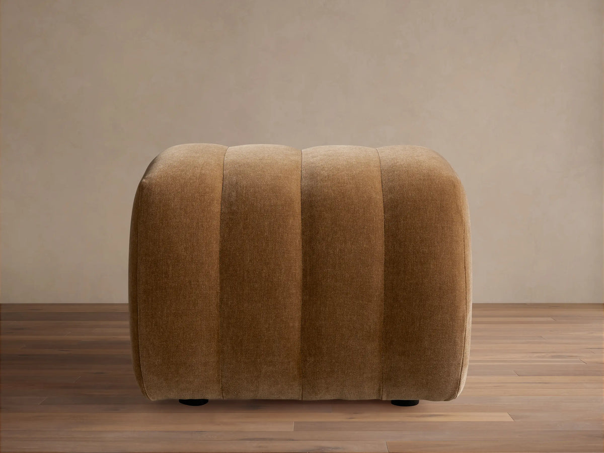 Warm brown Fresno Channel-Stitched Ottoman, a rounded, upholstered piece with vertical channels, on a wooden floor.