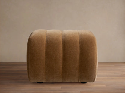 Warm brown Fresno Channel-Stitched Ottoman, a rounded, upholstered piece with vertical channels, on a wooden floor.