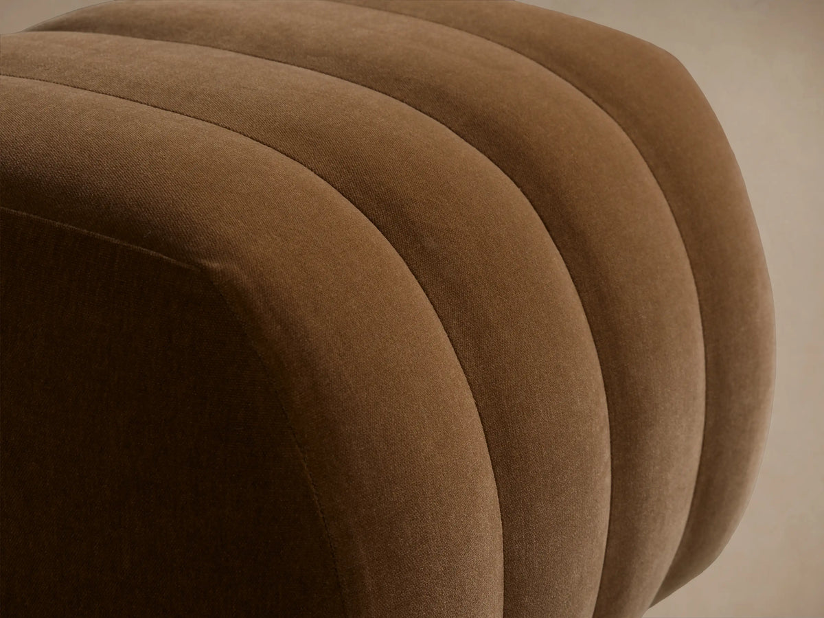 Warm brown Fresno Channel-Stitched Ottoman, a rounded, upholstered piece with vertical channels, on a wooden floor.