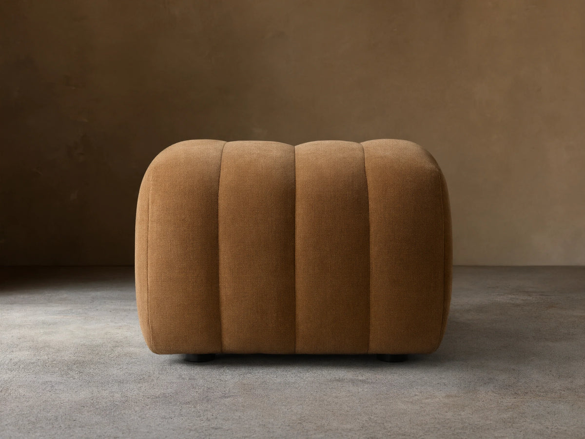 Warm brown Fresno Channel-Stitched Ottoman, a rounded, upholstered piece with vertical channels, on a wooden floor.