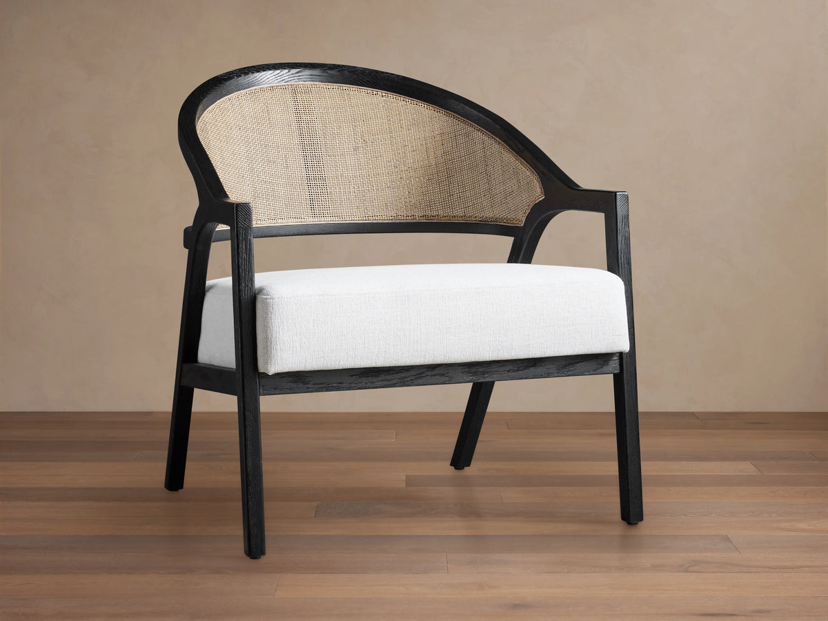 Aimee Chair: black oak frame, curved rattan cane back, and white Nomad Snow upholstered seat on a wood floor.