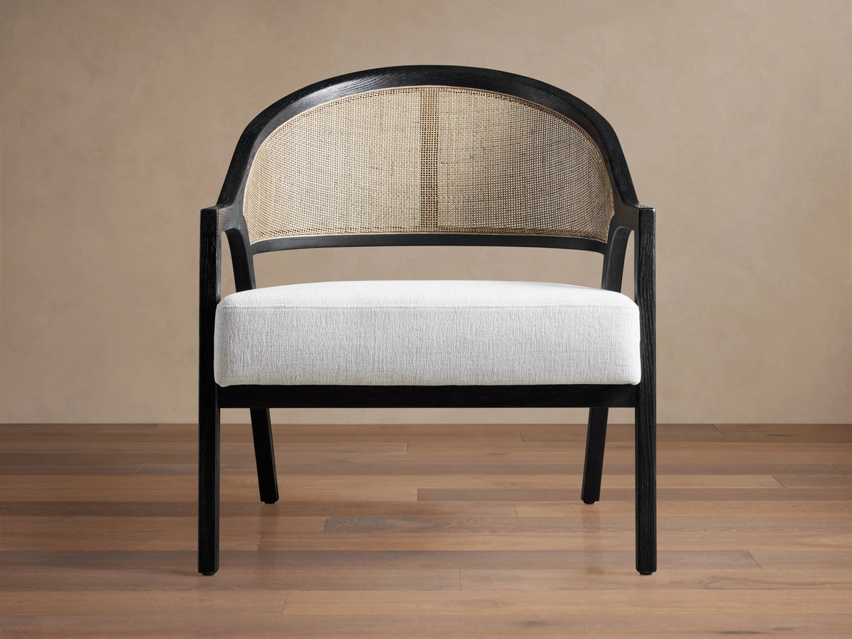 Aimee Chair: black oak frame, curved rattan cane back, and white Nomad Snow upholstered seat on a wood floor.