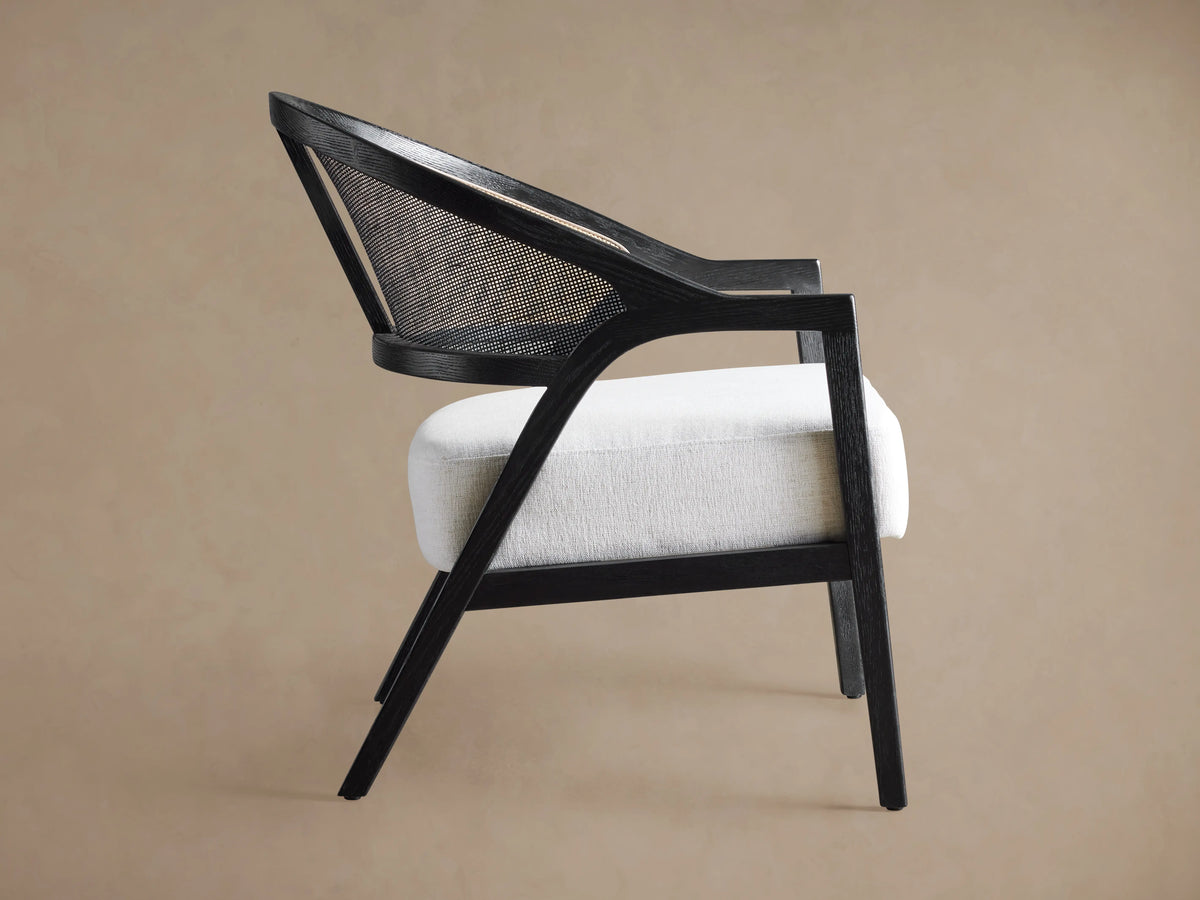 Aimee Chair: black oak frame, curved rattan cane back, light gray upholstered seat. Angled view on a light brown background.