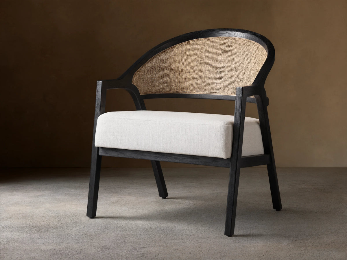 Aimee Chair: black oak frame, curved rattan cane back, and white Nomad Snow upholstered seat on a wood floor.
