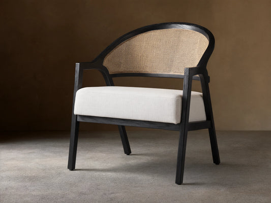 Aimee Chair: black oak frame, curved rattan cane back, and white Nomad Snow upholstered seat on a wood floor.