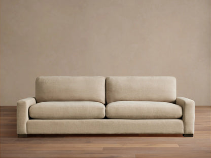 A beige Remington Sofa with plush cushions and a minimalist design sits on a wooden floor against a neutral wall.