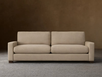 A beige Remington Sofa with plush cushions and a minimalist design sits on a wooden floor against a neutral wall.