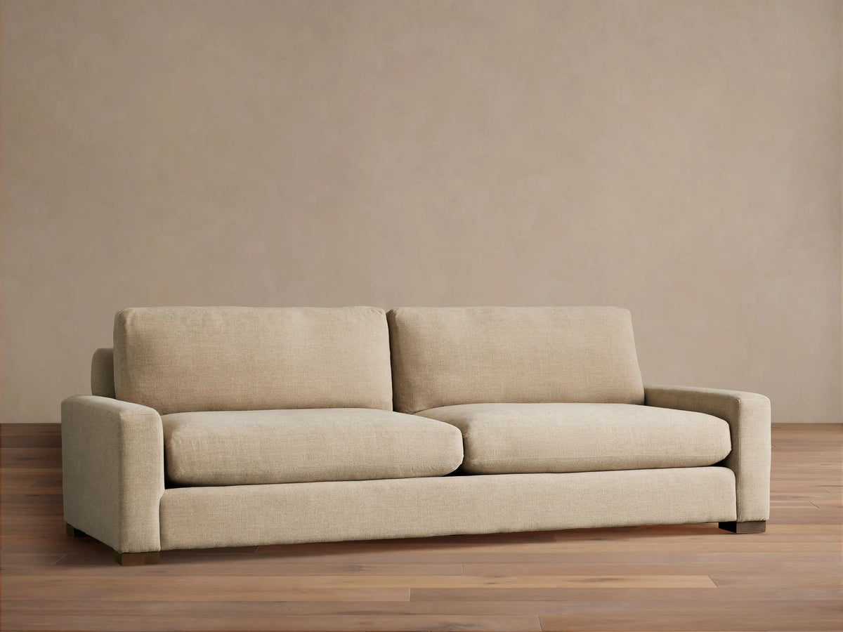 A beige Remington Sofa with plush cushions and a minimalist design sits on a wooden floor against a neutral wall.