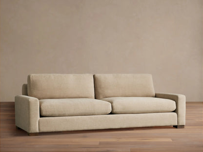 A beige Remington Sofa with plush cushions and a minimalist design sits on a wooden floor against a neutral wall.