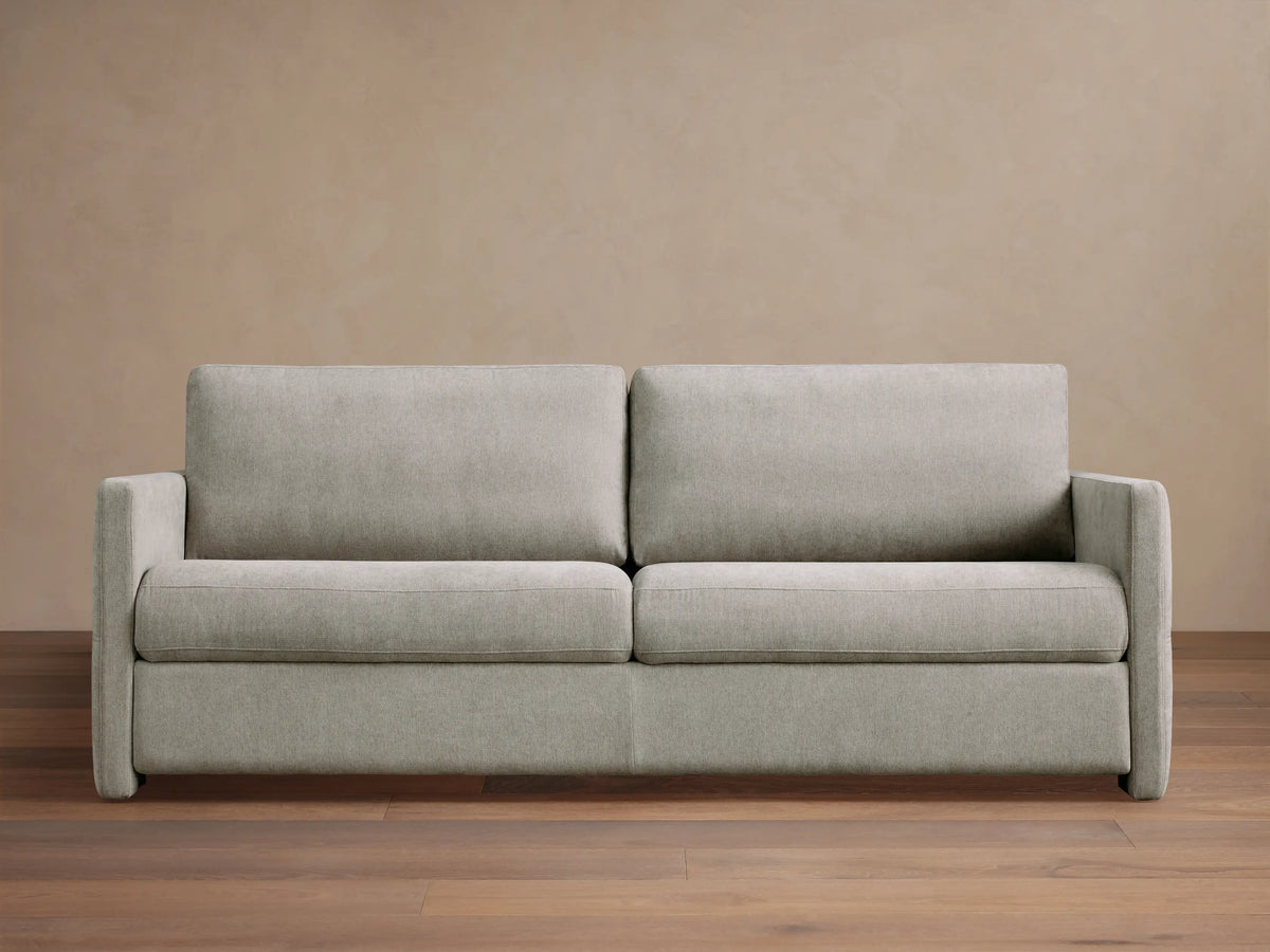 Reece Sleeper Sofa in light gray fabric, modern two-seater design with square arms, on a wooden floor against a tan wall.