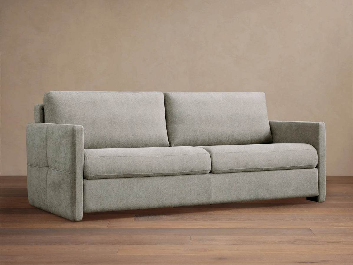 Reece Sleeper Sofa in light gray fabric, modern two-seater design with square arms, on a wooden floor against a tan wall.