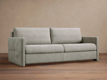 Reece Sleeper Sofa in light gray fabric, modern two-seater design with square arms, on a wooden floor against a tan wall.