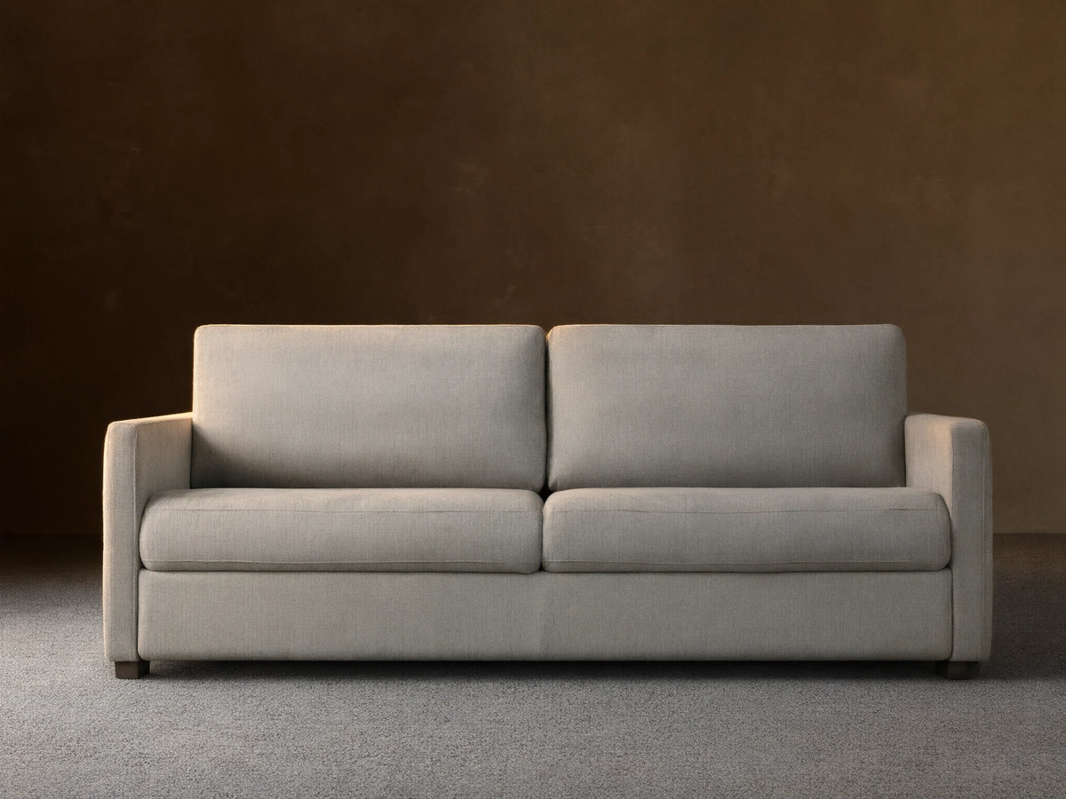 Reece Sleeper Sofa in light gray fabric, modern two-seater design with square arms, on a wooden floor against a tan wall.