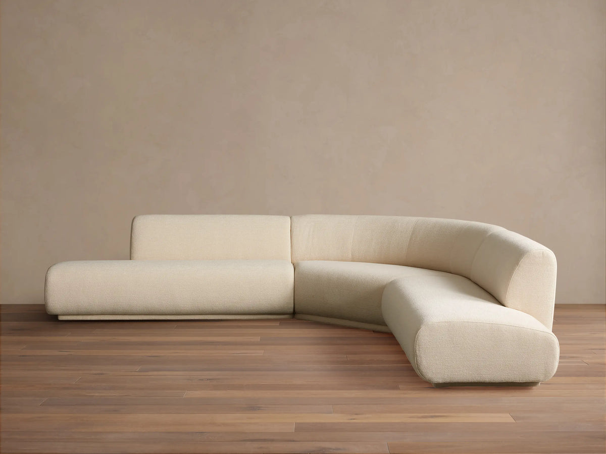 Cream Gianni Three-Piece Curved Bumper Sectional on a wooden floor against a tan wall, showing its elegant, modern design.