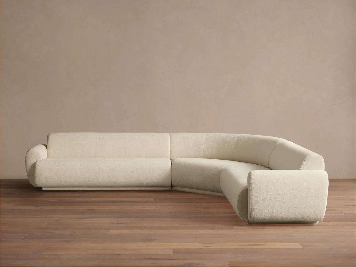Gianni Three-Piece Corner Sectional. Off-white curved L-shaped sofa with soft texture on a wooden floor, against a beige wall.
