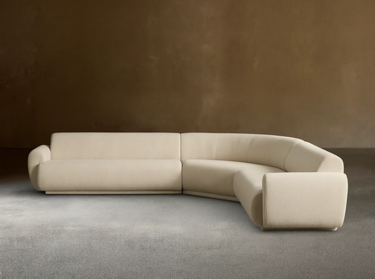 Gianni Three-Piece Corner Sectional. Off-white curved L-shaped sofa with soft texture on a wooden floor, against a beige wall.