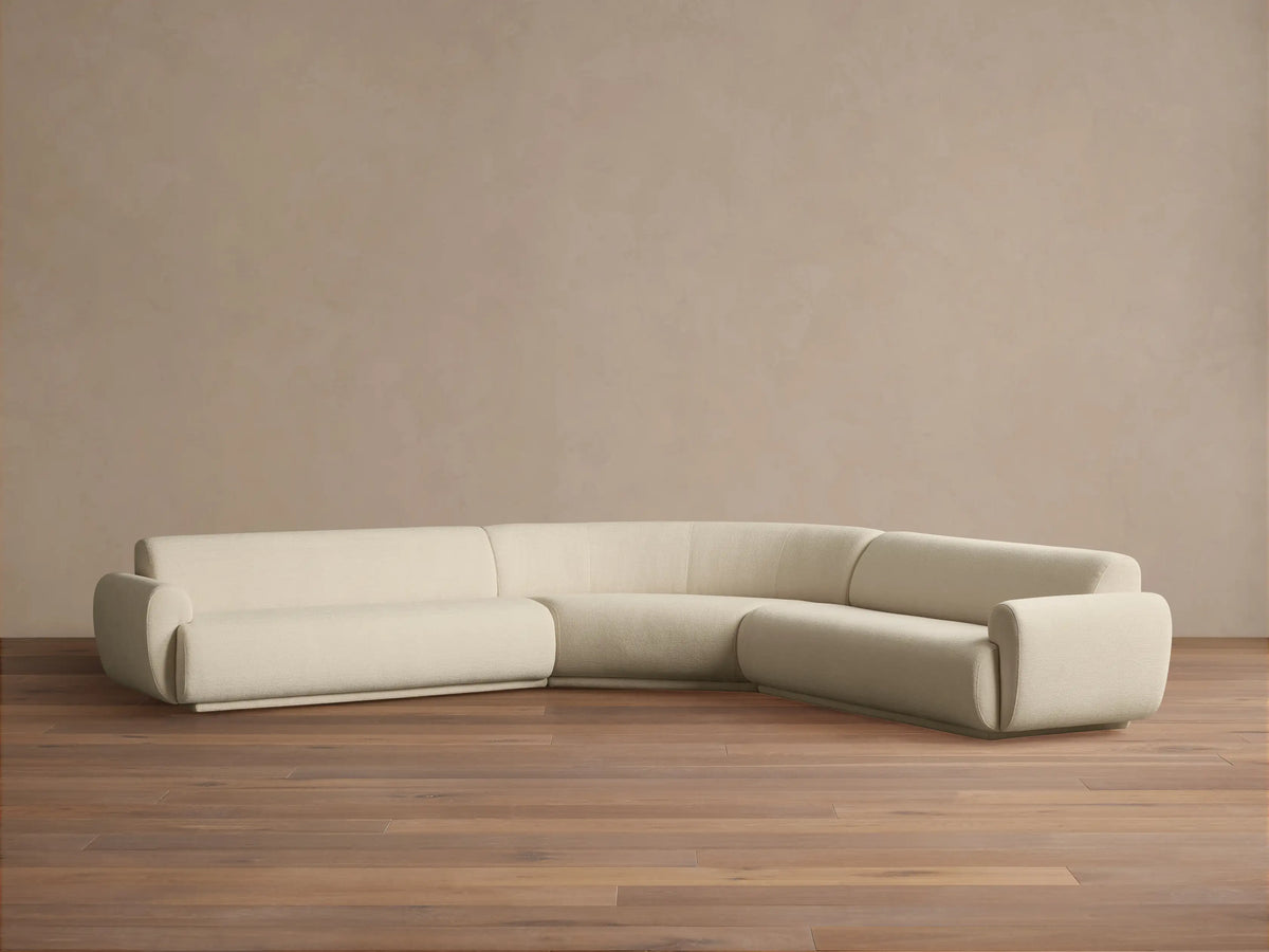 Gianni Three-Piece Corner Sectional. Off-white curved L-shaped sofa with soft texture on a wooden floor, against a beige wall.