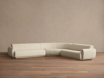 Gianni Three-Piece Corner Sectional. Off-white curved L-shaped sofa with soft texture on a wooden floor, against a beige wall.