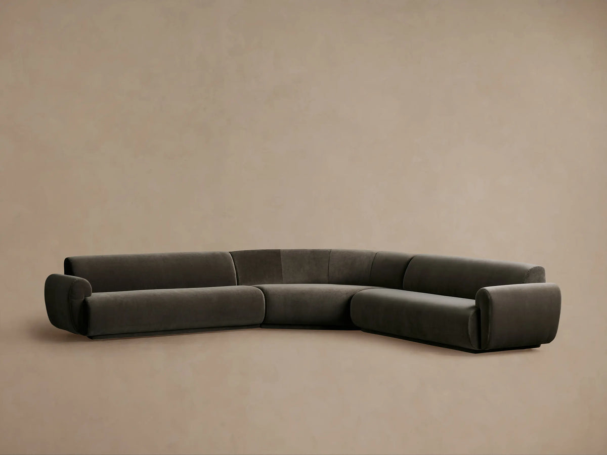 Gianni Three-Piece Corner Sectional in dark grey fabric, featuring a curved corner, on a wooden floor against a beige wall.