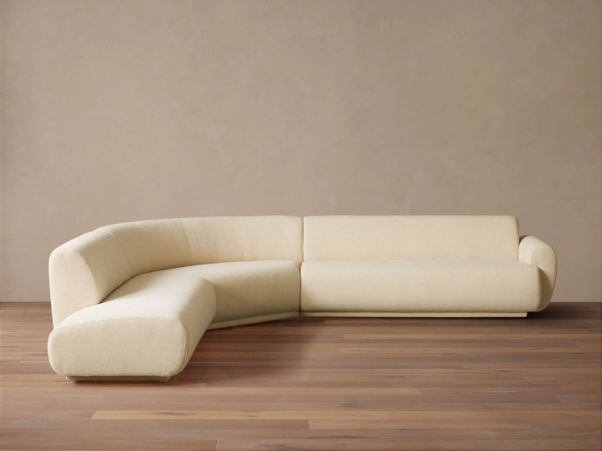 Gianni Three-Piece Bumper Sectional in cream, L-shaped with a curved left end, on a wood floor against a beige wall.