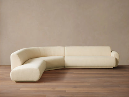 Gianni Three-Piece Bumper Sectional in cream, L-shaped with a curved left end, on a wood floor against a beige wall.