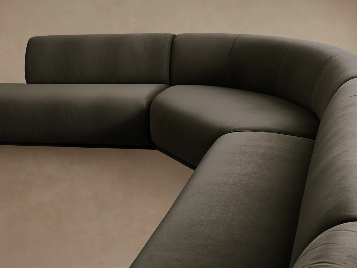 Gianni Five-Piece Curved Bumper Sectional in dark olive green velvet, on a wooden floor against a beige wall.