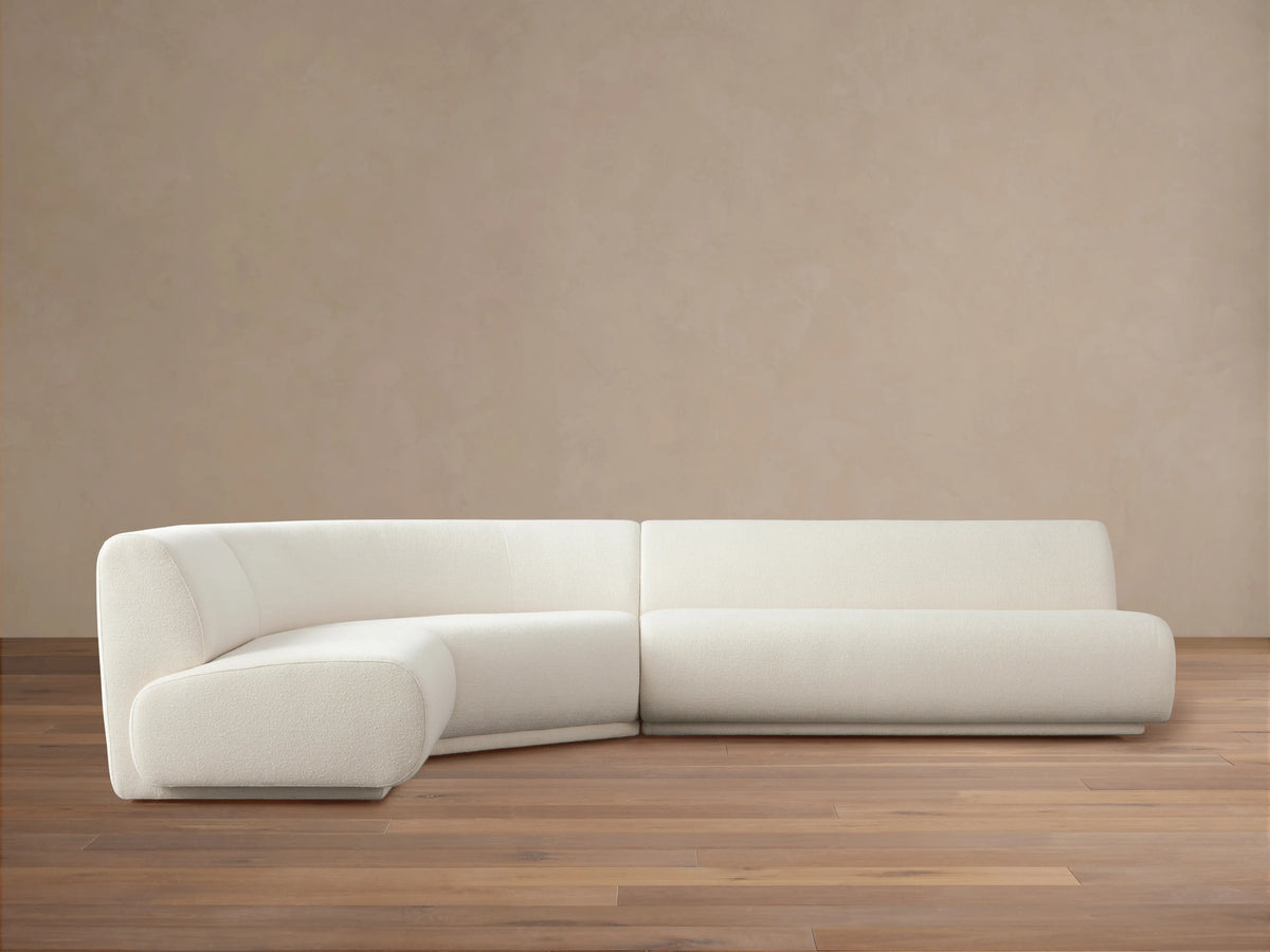 Gianni Three-Piece Sectional in off-white fabric, positioned on a wooden floor with a neutral beige background. Sectional features a curved left piece and long right piece.