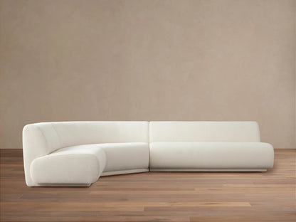 Gianni Three-Piece Sectional in off-white fabric, positioned on a wooden floor with a neutral beige background. Sectional features a curved left piece and long right piece.