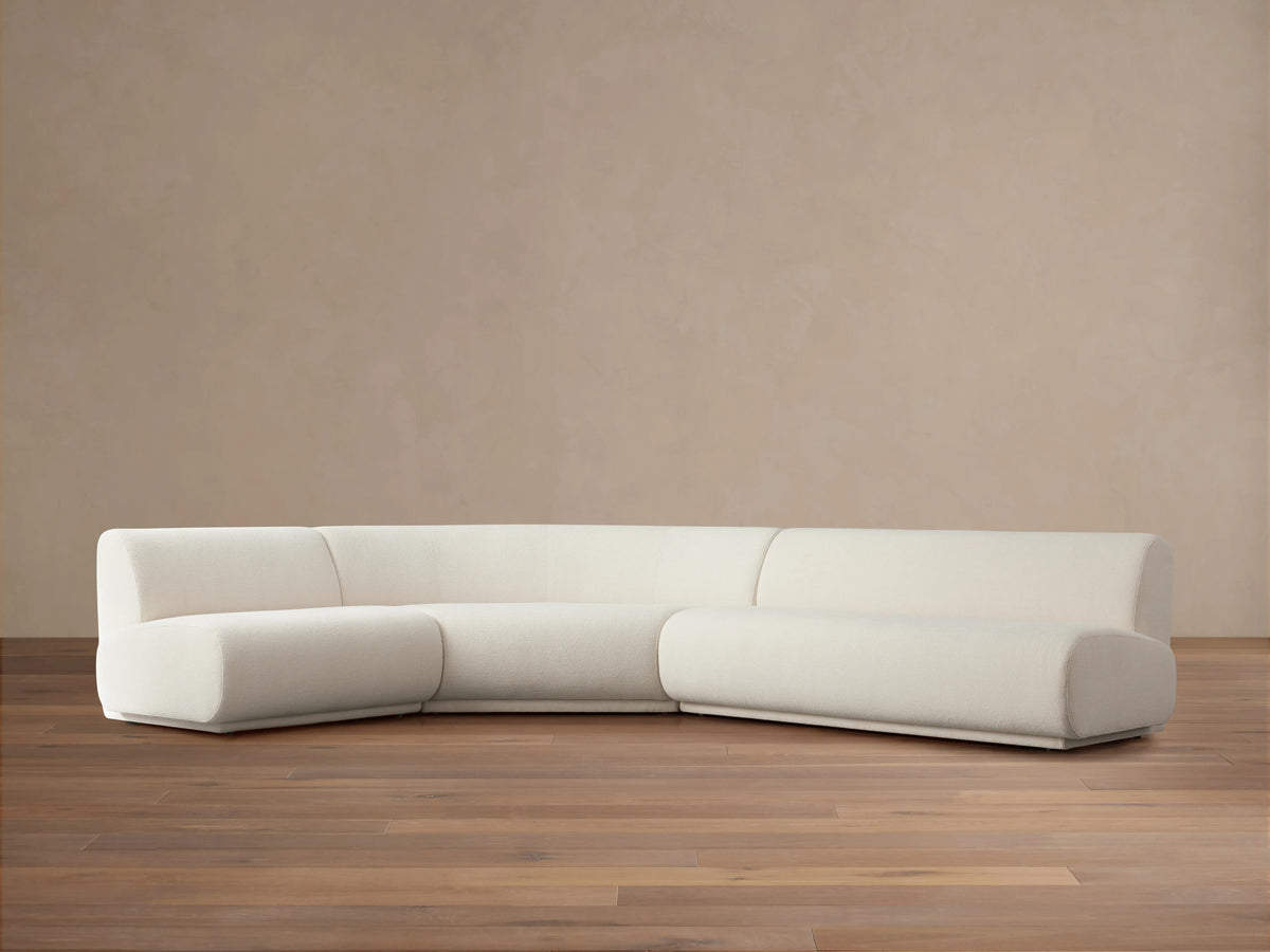 Gianni Three-Piece Sectional in off-white fabric, positioned on a wooden floor with a neutral beige background. Sectional features a curved left piece and long right piece.